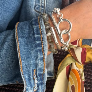 May include: A wrist featuring a collection of bracelets. A thick silver chain bracelet with a large clasp is worn alongside a silver bar bracelet and a beaded bracelet. The person is wearing a denim jacket and a patterned scarf.