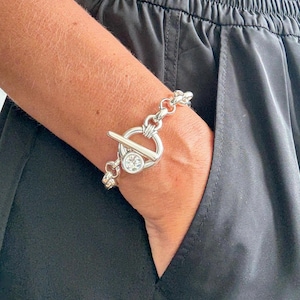 Puede incluir: Una pulsera plateada con cierre de palanca y una pieza central circular con una piedra transparente. La pulsera se lleva en la muñeca, con pantalones negros en el fondo.