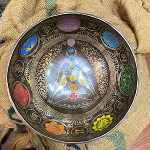 May include: A black singing bowl with gold and colorful designs. The bowl features a stylized figure sitting in a meditative pose with seven chakras depicted in different colors. The bowl is surrounded by symbols and designs.