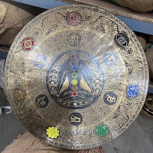 May include: A large, round, brass gong with intricate floral designs and a central image of a seated figure with seven chakras. The gong is decorated with colorful symbols and Sanskrit text.