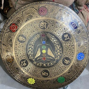 May include: A large, round, black metal gong with gold detailing. The gong features a central image of a meditating figure with seven chakras represented by colored circles. The gong is decorated with intricate floral patterns and Sanskrit symbols.
