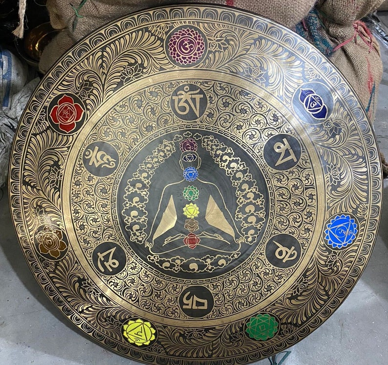 May include: A large, round, black metal gong with gold detailing. The gong features a central image of a meditating figure with seven chakras represented by colored circles. The gong is decorated with intricate floral patterns and Sanskrit symbols.