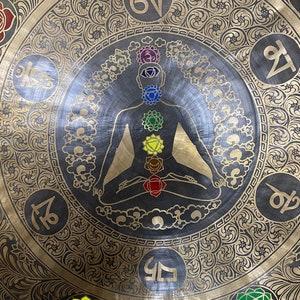 May include: A large, round, brass gong with intricate floral designs and a central image of a person in a meditative pose with seven colored chakras. The gong has Sanskrit symbols around the edge.