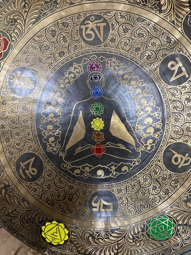 May include: A large, round, brass gong with intricate floral designs and a central image of a person in a meditative pose with seven colored chakras. The gong has Sanskrit symbols around the edge.