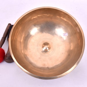 Puede incluir: Un cuenco de canto dorado grande con un mazo de madera y un batidor de fieltro rojo. El cuenco tiene una superficie lisa y pulida y una pequeña protuberancia elevada en el centro.