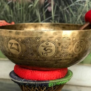 May include: A large, antique-looking, brass singing bowl with intricate engravings of symbols and a red felt mallet. The bowl is resting on a red and green cushion.