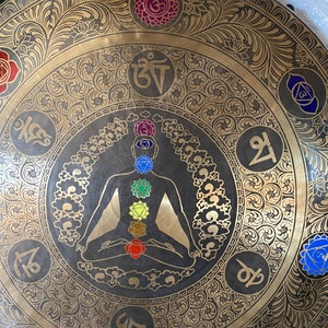 May include: A large, round, brass gong with a black silhouette of a person in a meditative pose. The gong is decorated with intricate floral patterns and seven colorful chakras, each with a Sanskrit symbol.