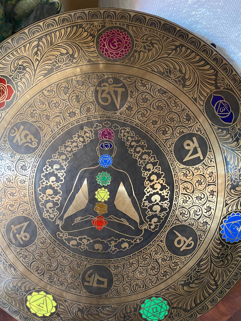 May include: A large, round, brass gong with a black silhouette of a person in a meditative pose. The gong is decorated with intricate floral patterns and seven colorful chakras, each with a Sanskrit symbol.