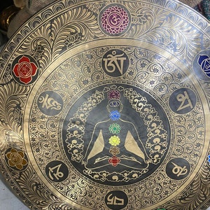 May include: A large, round, brass gong with intricate floral designs and a central image of a meditating figure with seven chakras. The gong has a black background and gold accents. The chakras are represented by colored circles, and the figure is surrounded by Sanskrit symbols.
