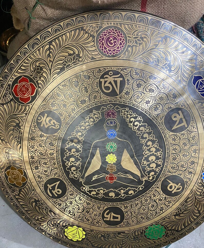 May include: A large, round, brass gong with intricate floral designs and a central image of a meditating figure with seven chakras. The gong has a black background and gold accents. The chakras are represented by colored circles, and the figure is surrounded by Sanskrit symbols.