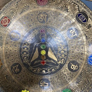 May include: A large, round, brass gong with intricate gold designs and a central image of a person in a meditative pose. The gong has seven colored circles around the person, representing the seven chakras. The gong also has various Sanskrit symbols around the edge.