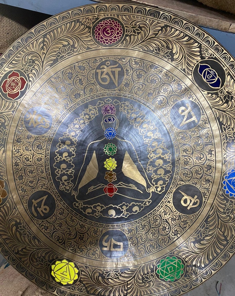 May include: A large, round, brass gong with intricate gold designs and a central image of a person in a meditative pose. The gong has seven colored circles around the person, representing the seven chakras. The gong also has various Sanskrit symbols around the edge.