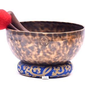 Puede incluir: Un cuenco de canto grande de aspecto antiguo con un acabado martillado y una pátina marrón oscura. El cuenco descansa sobre un cojín con estampado azul y dorado. Un mazo de fieltro rojo descansa en el borde del cuenco.