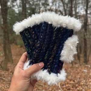 May include: A hand-knitted, dark blue fingerless glove with a textured pattern. The glove is trimmed with fluffy white faux fur. The yarn has flecks of green, yellow, and brown. The background is a blurred outdoor scene.