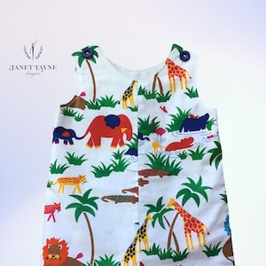 Puede incluir: Romper blanco sin mangas con estampado de animales de la selva. El estampado presenta elefantes, jirafas, leones, hipopótamos, cocodrilos y palmeras. El romper tiene botones azules en las tiras.