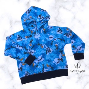May include: A blue hooded sweatshirt featuring a pattern of Olaf from Frozen and snowflakes. The cuffs and hem are black. The sweatshirt has a drawstring hood and long sleeves. The brand name "Janet Tayne Designs" is visible in the lower right corner.