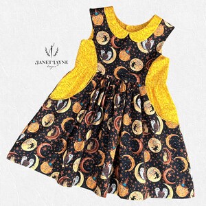 Könnte beinhalten: Ein schwarzes Kleid mit gelbem Kragen und Taschenakzenten. Das Kleid hat einen Aufdruck von Monden, Eulen und Kürbissen. Der Text "Janet Tayne designs" ist auf der linken Seite zu sehen.