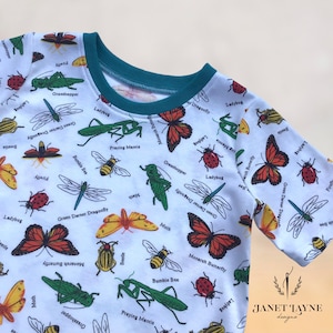May include: White t-shirt with a colorful insect print. The shirt features a variety of insects, including butterflies, dragonflies, ladybugs, and praying mantises. The text on the shirt includes the names of the insects, such as "Monarch Butterfly", "Green Darner Dragonfly", and "Praying Mantis".