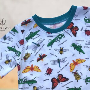 May include: White t-shirt with a teal neckband and a repeating pattern of various insects, including ladybugs, bumblebees, praying mantises, dragonflies, and moths. The shirt has the text "Janet Jayne Designs" in the top left corner.
