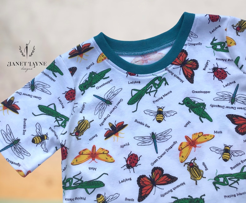 May include: White t-shirt with a teal neckband and a repeating pattern of various insects, including ladybugs, bumblebees, praying mantises, dragonflies, and moths. The shirt has the text "Janet Jayne Designs" in the top left corner.