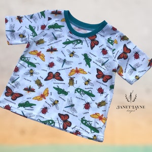 May include: White short-sleeved t-shirt with a colorful insect print. The print features a variety of insects, including butterflies, dragonflies, ladybugs, and grasshoppers. The text "Janet Jayne Designs" is printed on the bottom right corner of the shirt.