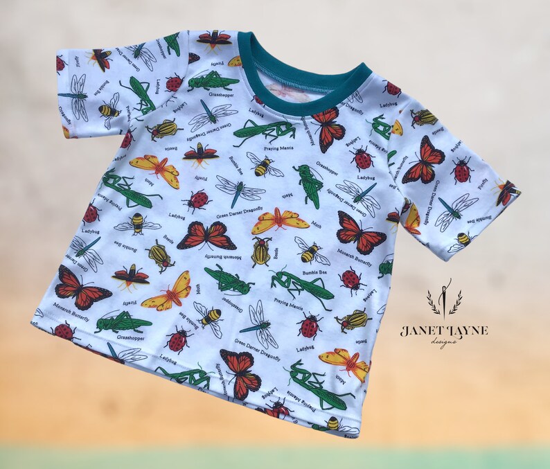 May include: White short-sleeved t-shirt with a colorful insect print. The print features a variety of insects, including butterflies, dragonflies, ladybugs, and grasshoppers. The text "Janet Jayne Designs" is printed on the bottom right corner of the shirt.