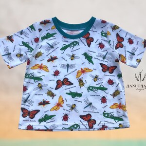 May include: White short-sleeved t-shirt with a colorful insect print. The print features butterflies, ladybugs, dragonflies, bees, and praying mantises. The shirt has a teal colored neckline.