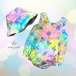 May include: A colorful baby romper and matching sun hat set. The fabric features a tie-dye effect in shades of pink, yellow, blue, and green, with a repeating blue bee pattern. The romper has snap closures and the hat has a brim.
