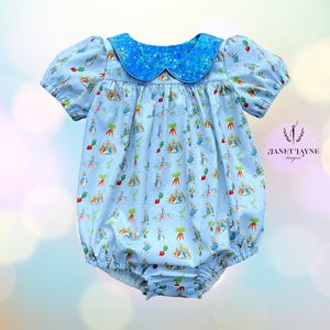 May include: A light blue baby romper with a Peter Rabbit print, featuring a contrasting blue floral collar and short puff sleeves. The romper has a gathered waist and snap closure. The text "JANET TAYNE designs" is visible.