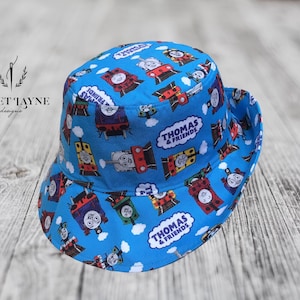 May include: A blue bucket hat with a Thomas & Friends print featuring various characters from the show. The hat has a wide brim and a flat crown.
