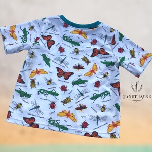 May include: White short-sleeved t-shirt with a colorful insect print. The print features ladybugs, butterflies, dragonflies, praying mantis, and other insects. The shirt has a teal blue collar.