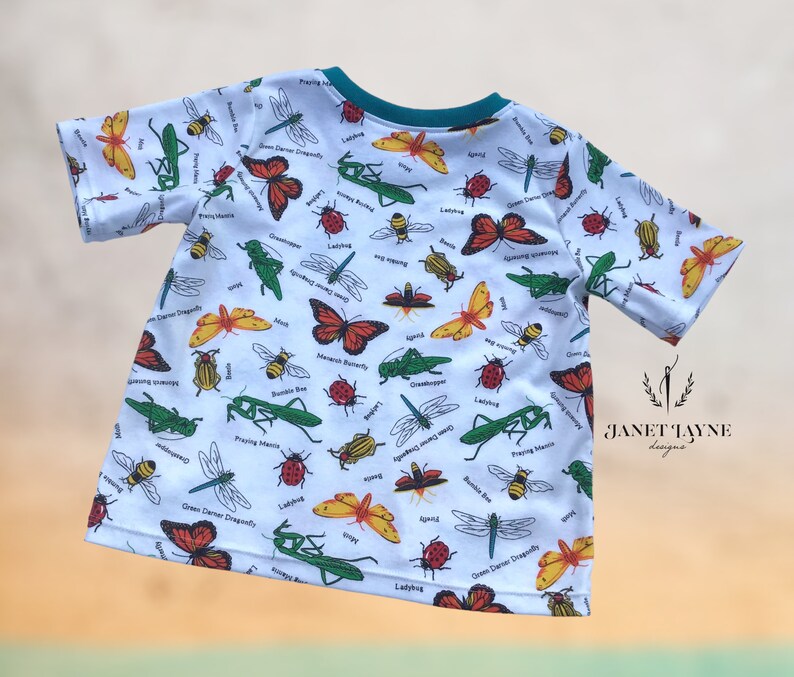 May include: White short-sleeved t-shirt with a colorful insect print. The print features ladybugs, butterflies, dragonflies, praying mantis, and other insects. The shirt has a teal blue collar.