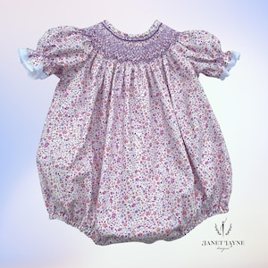 May include: A white baby bubble with a purple smocked bodice and a pink and purple floral print. The bubble has short puffed sleeves with white lace trim. The bubble is made by Janet Layne Designs.