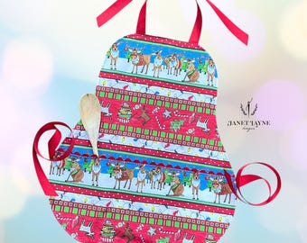 Puede incluir: Delantal de cocina rojo navideño con un diseño de renos y golosinas festivas. El delantal tiene un bolsillo y lazos de cinta roja. Una cuchara de madera está en el bolsillo. Diseñado por Janet Tayne Designs.