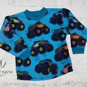 May include: A long-sleeved, turquoise fleece shirt featuring a repeating pattern of colorful monster trucks and star shapes. The shirt has a crew neck and cuffs in a darker teal color. The design is playful and suitable for children.