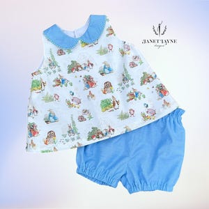May include: A two-piece baby outfit featuring a sleeveless top and matching shorts. The top has a light blue collar and is patterned with illustrations of rabbits and other characters. The shorts are a solid light blue color. The outfit is from Janet Tayne Designs.