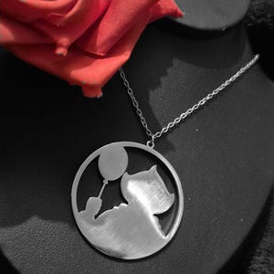 May include: A silver-colored pendant necklace featuring a circular design with a silhouette of a person holding a balloon and a cat. The necklace is displayed on a black surface, with a red rose in the background.