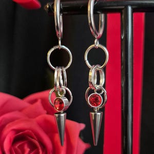 May include: A pair of silver earrings with red crystal accents. Each earring features a hoop with multiple smaller hoops and a dangling spike with a red crystal.