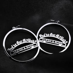 Op de afbeelding: Een paar zilveren oorringen. Elke oorring heeft de tekst "God he Got My Body" en "FLESH & BONE" in een gotisch lettertype. De oorbellen zijn cirkelvormig en hebben een glanzende afwerking.