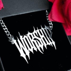 May include: A silver-colored necklace with the word "WORSHIP" in a bold, gothic font. The pendant is connected to a chain. The necklace is displayed in a black box, with a red rose in the background.
