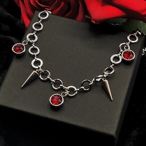 May include: A silver chain bracelet with red gemstone charms and silver spikes. The bracelet is on a black background.