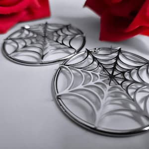 Könnte beinhalten: Ein Paar silberfarbene Creolen mit Spinnennetz-Design. Die runden Ohrringe weisen ein detailliertes Netzmuster auf. Roter Stoff ist im Hintergrund zu sehen und setzt einen Farbakzent in dem Bild.