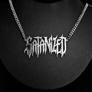 May include: Silver-toned necklace with the word "SATANIZED" in a gothic font. The pendant hangs from a curb chain. The necklace is displayed on a black bust, creating a stark contrast.