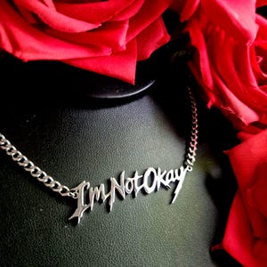 Puede incluir: Un collar plateado con la frase "I'm Not Okay" en una fuente gótica. El collar se exhibe sobre una superficie negra, con rosas rojas en el fondo.