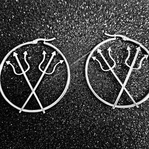 May include: A pair of silver hoop earrings, each with a trident motif inside the hoop. The tridents are crossed, forming a distinctive design. The earrings are set against a textured, dark background, creating a contrast.