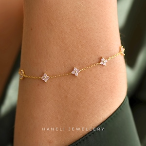 May include: A fine gold bracelet with tiny, four-petal flower charms, each with a pink gemstone centre. The bracelet is shown on an arm, with the brand name "HANELI JEWELLERY" visible at the bottom.