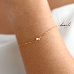 May include: A delicate gold chain bracelet featuring a small central cluster with a single round white opal and two tiny clear accent stones. The text haneli JEWELLERY EST 2020 is printed in black in the upper right corner of the image.