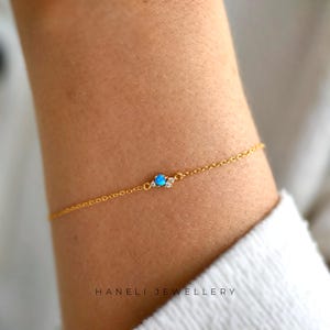 May include: A fine gold bracelet with a small, round, blue opal stone and two clear stones. The bracelet is attached to a delicate chain. The text "HANELI JEWELLERY" is printed in white at the bottom.
