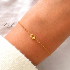 May include: A delicate gold chain bracelet featuring a central knot charm. The bracelet is thin and minimalist in construction. The text 'haneli JEWELLERY EST. 2020' is printed in black on the upper left side of the image, near the gold chain.