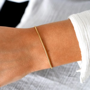 May include: A gold chain bracelet with a delicate, snake-like design. The bracelet is shown on a person's wrist.